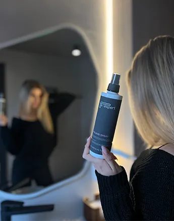 Жената държи спрей за коса пред огледалото, използвайки продукти за обем.